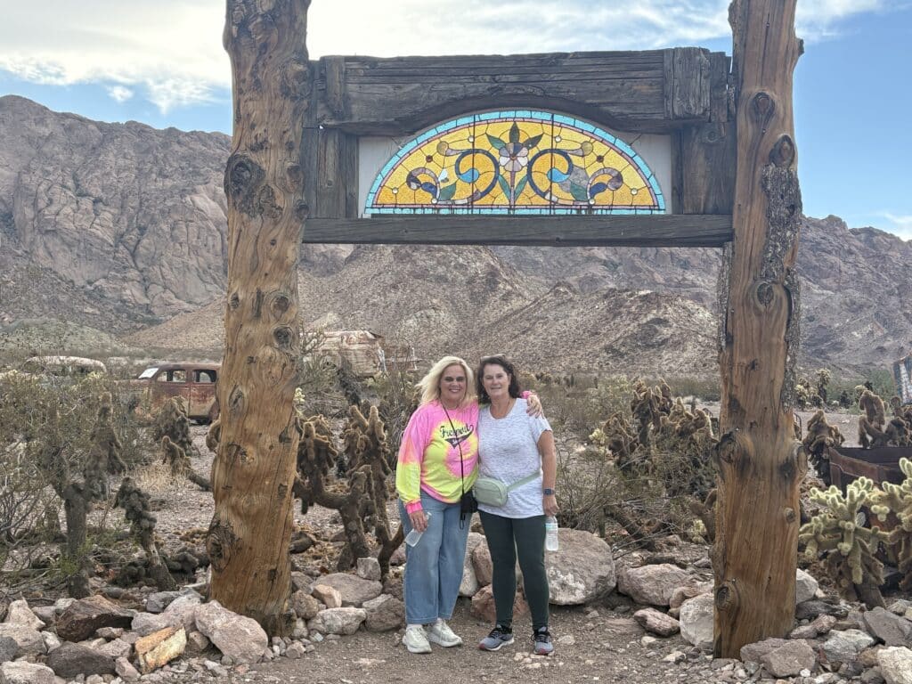 Two women smiling under a rustic arch with stained glass, desert plants, and Nevada mountains—perfect for Las Vegas sightseeing.