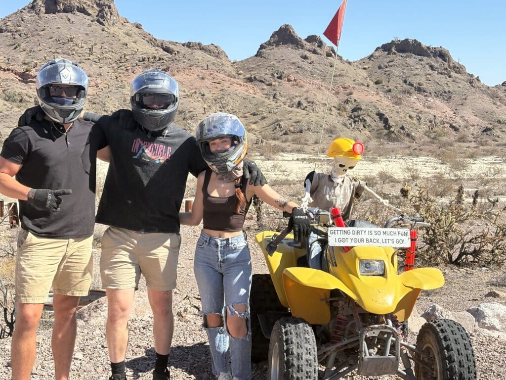 Adventurers in helmets pose by a yellow ATV with a skeleton rider, ready for Nevada desert off-road tours near Las Vegas.