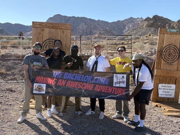 Six men with a banner, “The Bachelor Crew Having an Awesome Adventure,” pose by targets in Nevada near rocky mountains and clear sky.