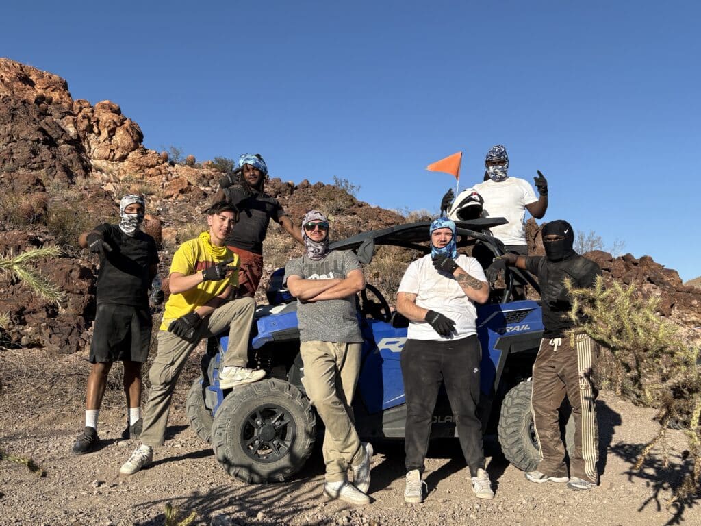 Group on ATV RZR tour poses by blue off-road vehicle near Las Vegas, Nevada desert rocks, cacti, and Colorado River views.