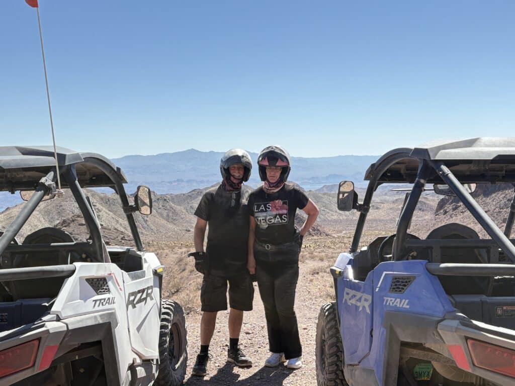 Two riders in black gear pose between ATVs on a Nevada mountain trail, blue sky and distant Las Vegas scenery in the background.