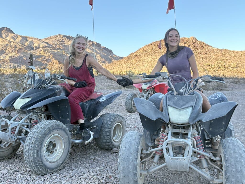 Two women enjoy an ATV tour near Las Vegas, Nevada, smiling and holding hands in the rocky desert with mountain views.