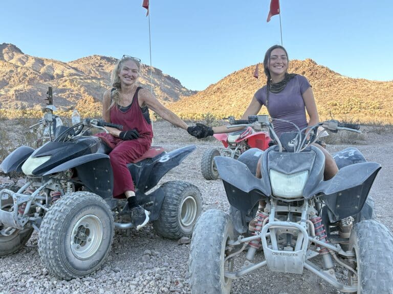 Two women enjoy an ATV tour near Las Vegas, Nevada, smiling and holding hands in the rocky desert with mountain views.