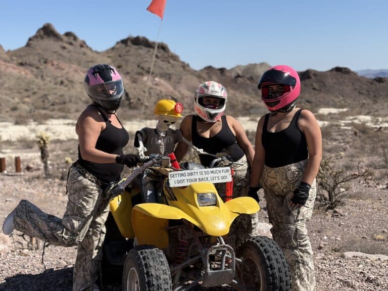 Three women in pink helmets pose by a yellow ATV near Las Vegas desert; skeleton rides ATV. Nevada ghost town ATV tour.