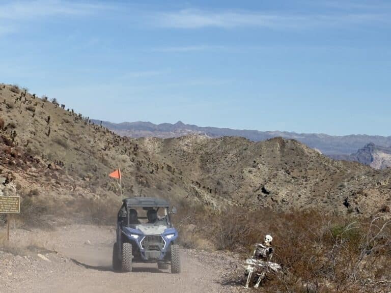 RZR off-road vehicle on a Nevada desert trail near Las Vegas; skeleton biker, sign, and rocky hills in the background.