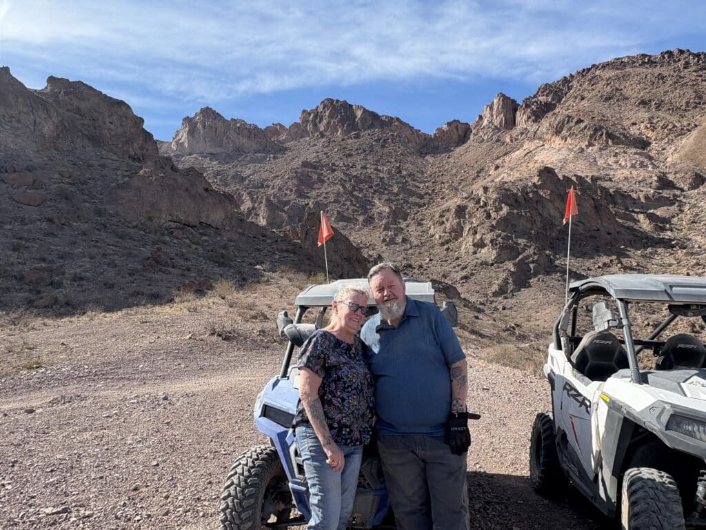 Smiling couple by two RZRs with red flags, desert terrain near Las Vegas, Nevada—perfect for ATV tours and off-road adventures.
