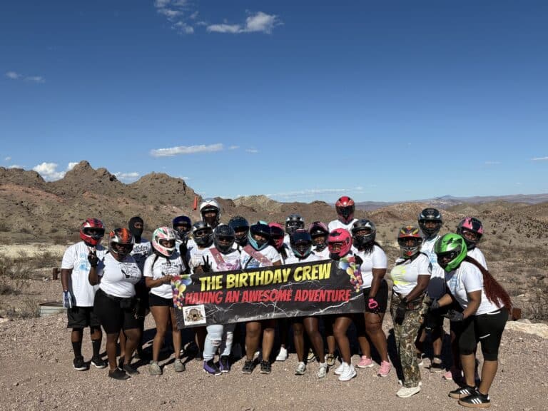 Friends in helmets and casual wear pose with a Birthday Crew banner during an ATV tour near the Colorado River in Nevada desert.