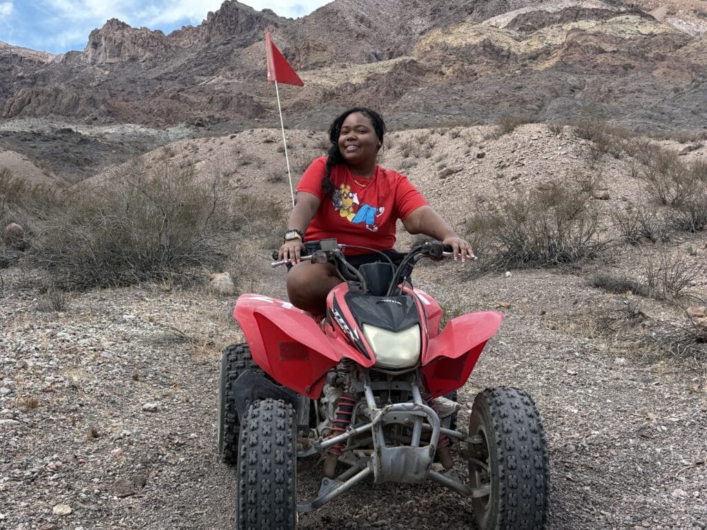 ATV tour in Nevada: person in red shirt rides red ATV on rocky desert terrain near dry bushes and mountains under cloudy sky.