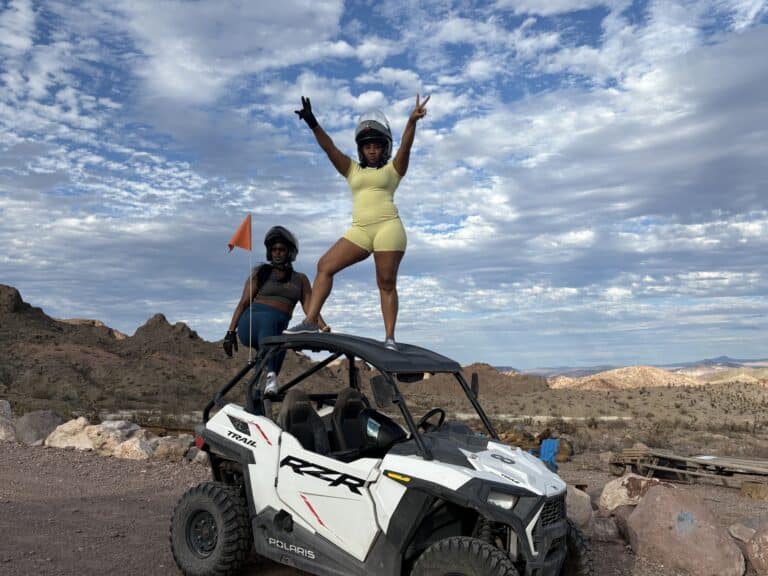 Adventurers in helmets pose on a white ATV during a Nevada desert RZR tour near Las Vegas, with mountains and clouds behind.