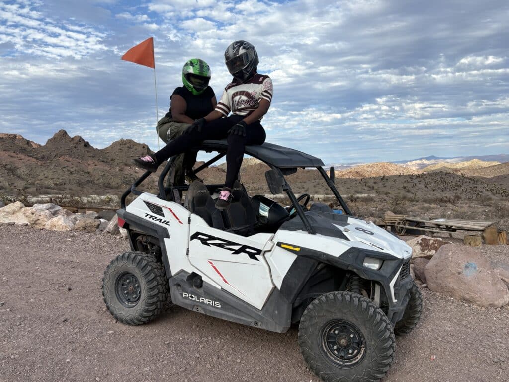Two in helmets on a white Polaris RZR in Nevada desert, orange flag flying—Las Vegas ATV tour near mountains and rocky trails.