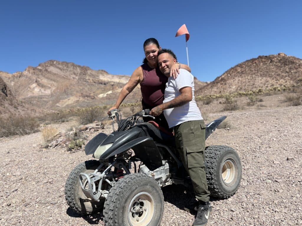Smiling friends on a black ATV in Nevada's rocky desert, mountains behind, enjoying an ATV tour near Las Vegas under blue skies.