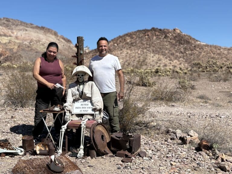 Two smiling tourists near a miner skeleton in a Nevada ghost town on an ATV tour, with desert and old mining gear in the background.