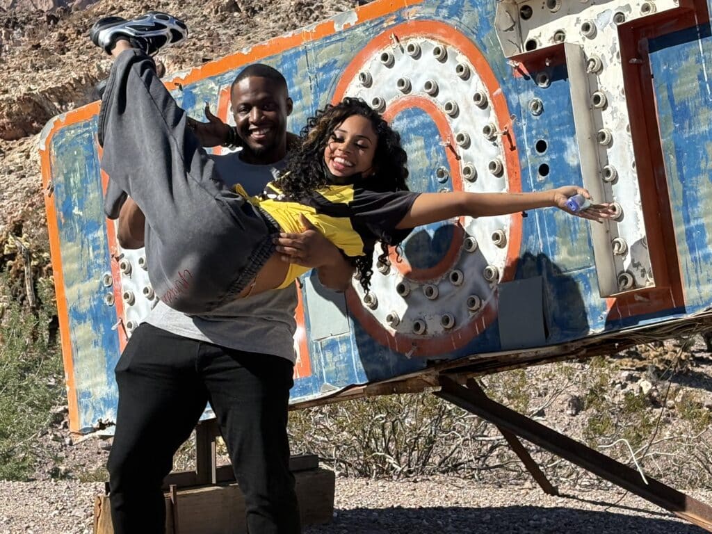 A man lifts a laughing woman by a vintage Las Vegas sign, with desert rocks and shrubs—perfect for Nevada Ghost Town sightseeing.