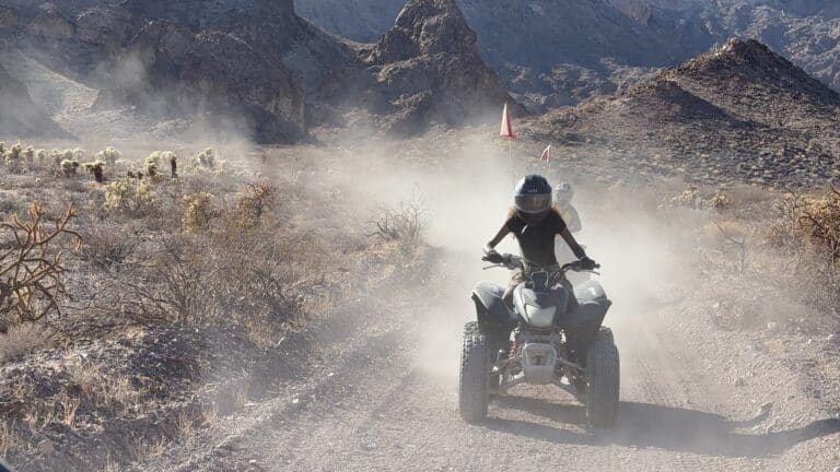 Rider in helmet drives ATV on a dusty Nevada desert trail near Las Vegas with rocky mountains and sparse vegetation.