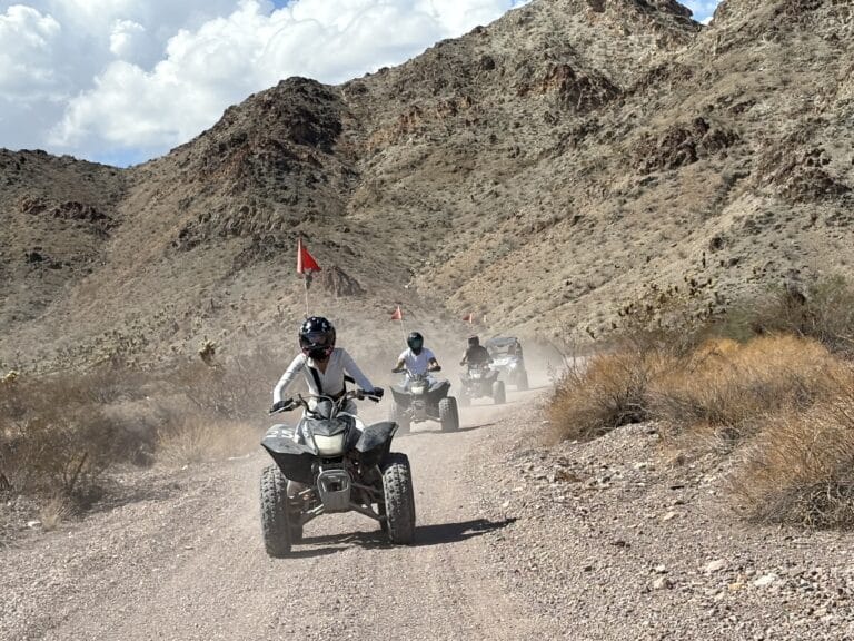 Adventurers ride ATVs on a dusty Nevada trail near Las Vegas with red flags, scenic mountains, and desert landscapes.