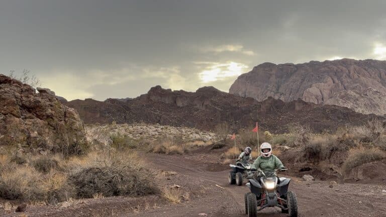 ATV riders explore a rocky Nevada desert trail near Las Vegas, with dry bushes, clouds, sunlight, and mountains in the background.