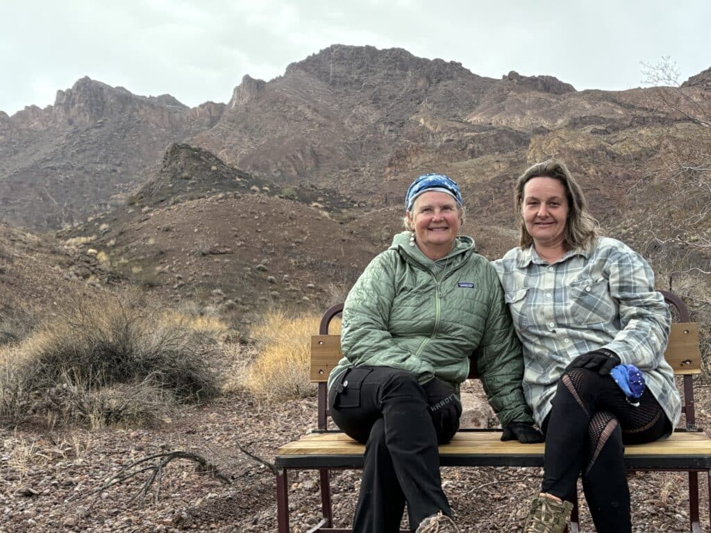 Two women in outdoor gear smile on a bench near rugged Nevada desert, with Las Vegas mountains, perfect for ATV or RZR rides.