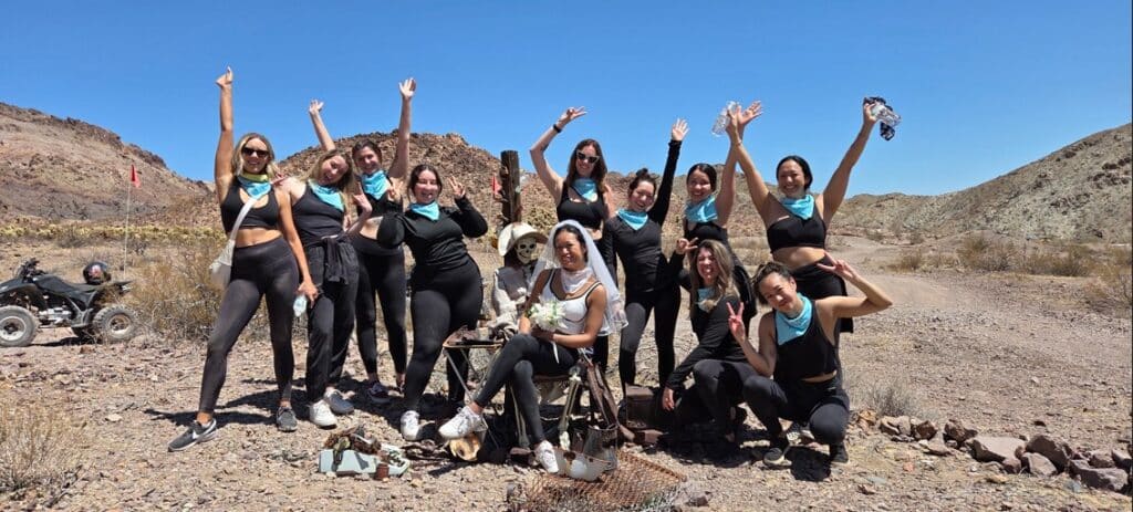 Women in black outfits and blue bandanas pose on a Las Vegas ATV desert tour near the Colorado River, ATV rider in background.