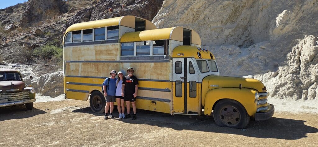 Smiling friends pose by a yellow vintage bus in Nevada near rocky hills, ready for Ghost Town sightseeing and ATV tours.