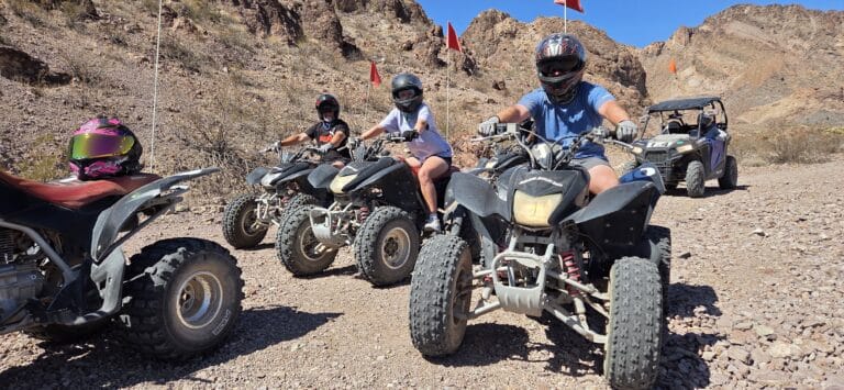 ATV riders in helmets explore a rocky Nevada desert trail near Las Vegas, with RZR in view and mountains in the background.