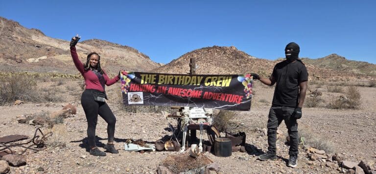 Two people pose in a Nevada desert near Las Vegas with a “Birthday Crew” banner, smiling after a Colorado River ATV adventure.