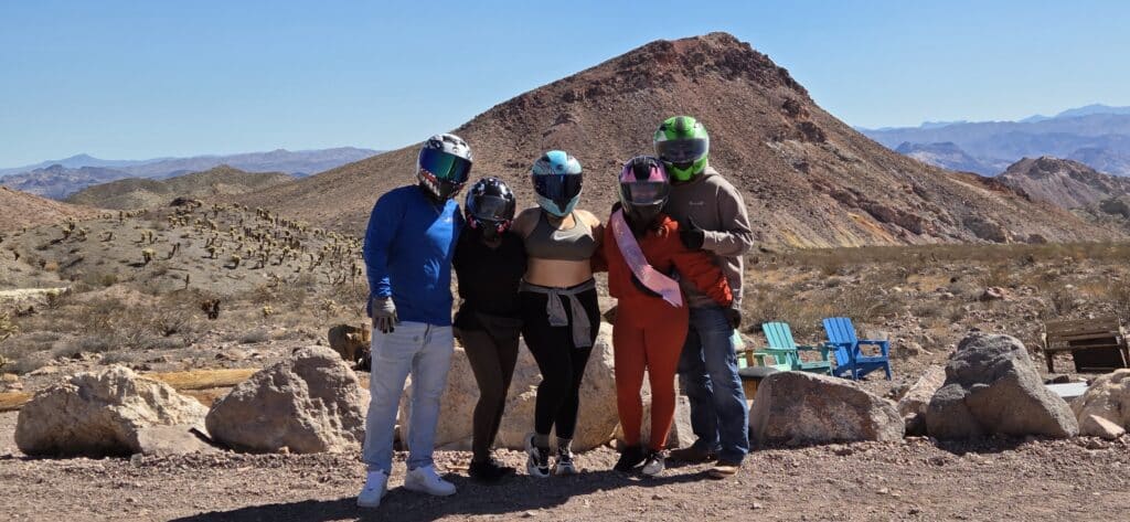 Group in helmets and casual wear enjoy Nevada desert near Las Vegas, with ATVs, blue chairs, rocks, and mountain views.