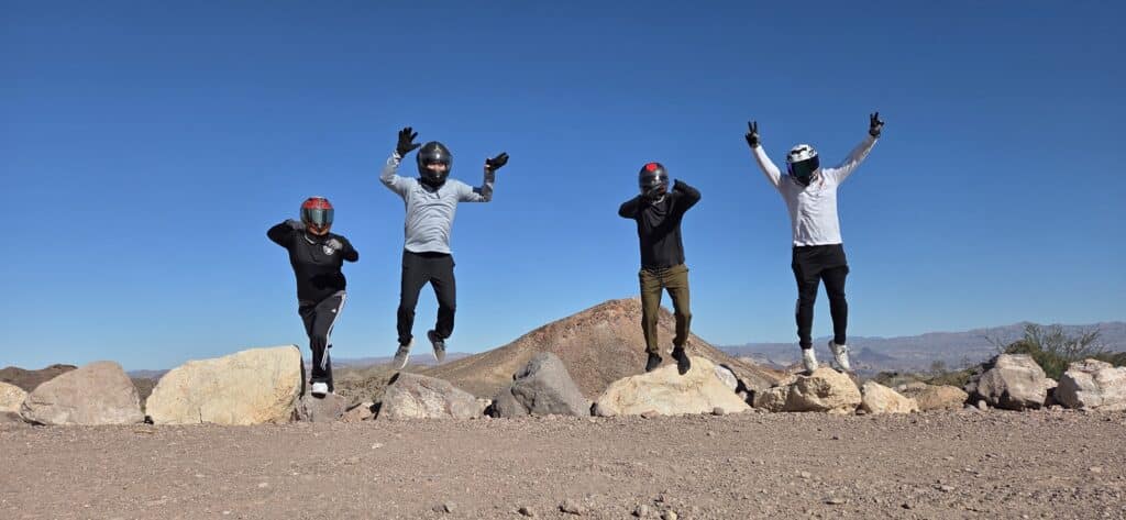 Four adventurers in helmets jump behind rocks on a Nevada desert ATV tour near Las Vegas under a vibrant blue sky.