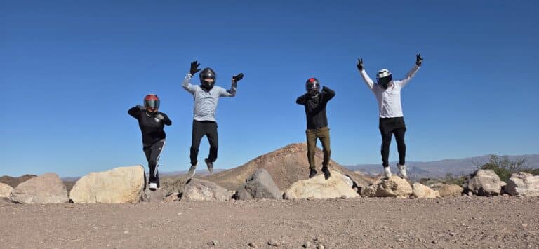 Four adventurers in helmets jump behind rocks on a Nevada desert ATV tour near Las Vegas under a vibrant blue sky.