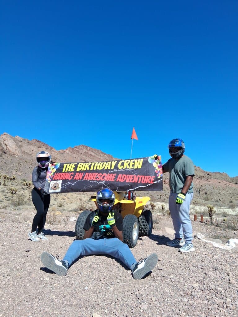 Three helmeted riders pose by an ATV in Nevada desert near Las Vegas, banner: THE BIRTHDAY CREW HAVING AN AWESOME ADVENTURE.