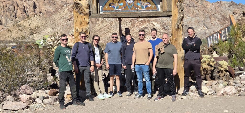 Group poses under a rustic arch with stained glass in Nevada desert—cacti, rocky hills, and Las Vegas Ghost Town sights behind.