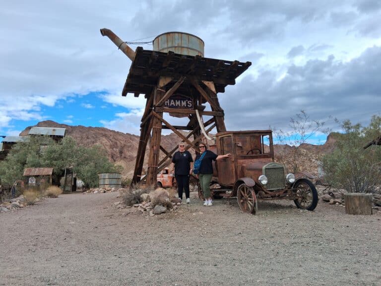 Two people pose by a rusty vintage car near HAMMS water tower in a Nevada ghost town, perfect for Las Vegas ATV and RZR tours.