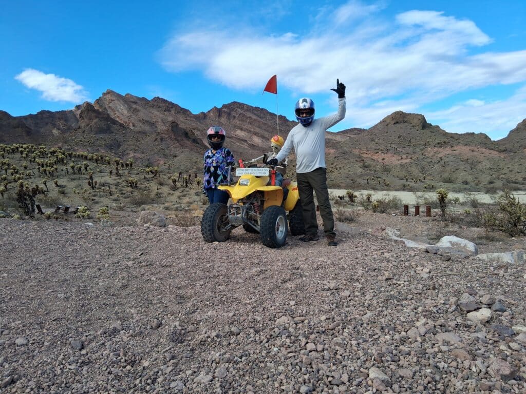 Two people in helmets by a yellow ATV on a Nevada desert ATV tour near Las Vegas, mountains and cacti under blue sky.