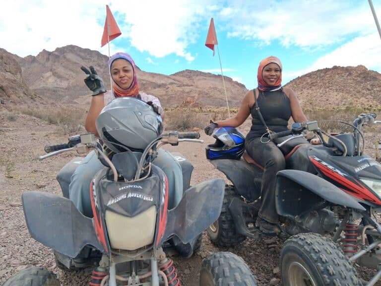 Two women on ATVs in Nevada desert, near Las Vegas, pose with peace sign during scenic ATV tour; mountains and blue sky behind.