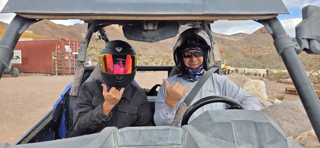 Adventure seekers enjoy a Las Vegas RZR off-road ride, flashing shaka signs amid Nevada desert terrain and a red shipping container.
