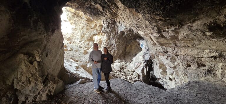 Two people stand in a rocky Nevada cave near the Colorado River, sunlight streaming in—perfect for RZR off-road tour adventures.