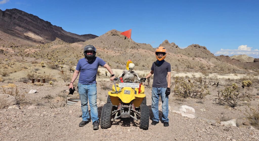 Two people in helmets pose by a yellow ATV with mannequin near Nevada mountains, ideal for Las Vegas ATV tours and Ghost Town rides.