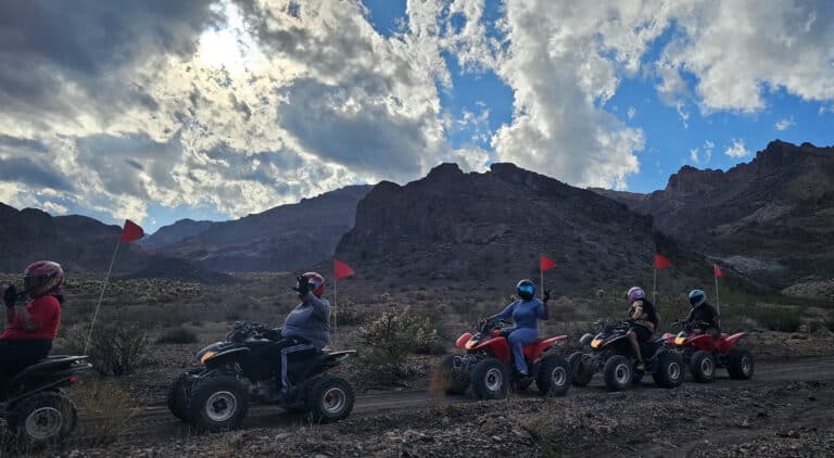 ATV riders in helmets explore a rocky Nevada desert trail near Las Vegas, with red flags, mountains, and dramatic clouds in view.
