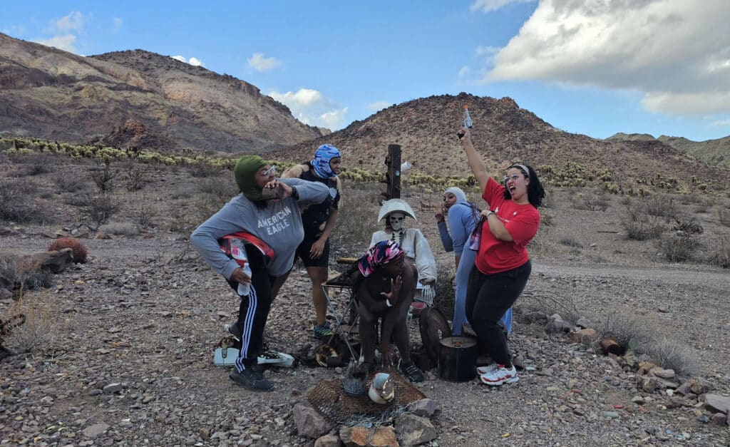 Group poses playfully near a rustic statue in Nevada desert, wearing bike gear; selfie during Las Vegas Ghost Town sightseeing.
