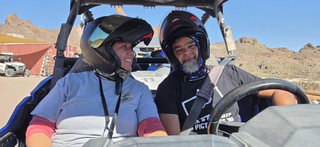 Two riders in helmets enjoy a Las Vegas RZR off-road tour, smiling with Nevada hills and a red building beneath clear blue skies.