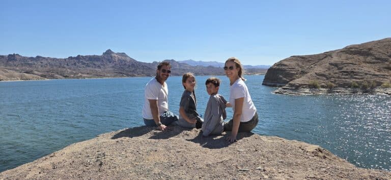 Family of four smiles on a rocky ledge near the Colorado River, Nevada, mountains and blue lake in view, perfect for ATV tours.
