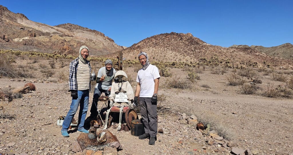 Three people pose by a dressed skeleton and sign in Nevada desert near Las Vegas, perfect for ghost town sightseeing adventures.