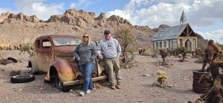 Couple poses by a rusty car in a Nevada desert ghost town near Las Vegas, old church and rocky hills in the background.
