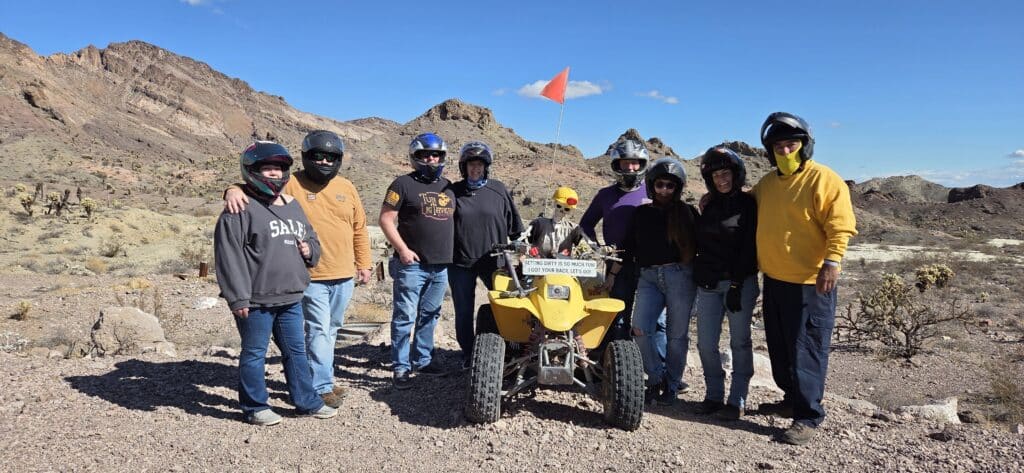 Group on Nevada ATV tour near Las Vegas, posing with yellow ATV and orange flag, mountains and desert sky in the background.