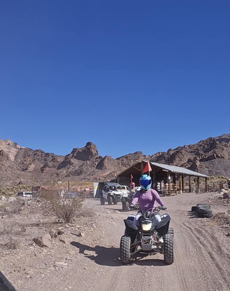 ATV rider in blue helmet explores Nevada desert near Las Vegas, with Ghost Town sights and Colorado River ATV tour vehicles behind.