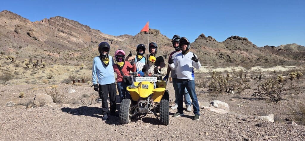Six adventurers in helmets pose by a yellow ATV on a Nevada desert tour near Las Vegas with mountains under clear blue skies.