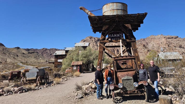 Group by rusty vintage car, HAMMS BEER water tower in Nevada ghost town near Las Vegas—ideal for ATV tours and sightseeing.