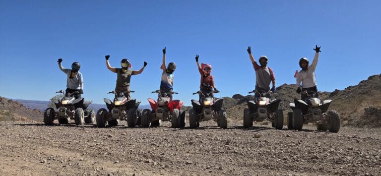 Group on ATVs raise arms during a Nevada ATV tour near Las Vegas, riding rocky desert trails under a clear sky.