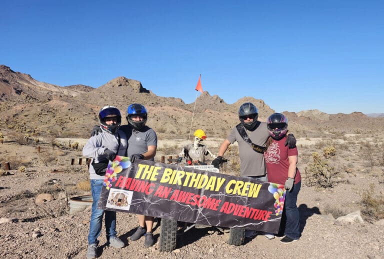 Birthday crew in helmets with banner, celebrating in Nevada desert near Las Vegas after an RZR ATV tour, mountains behind.