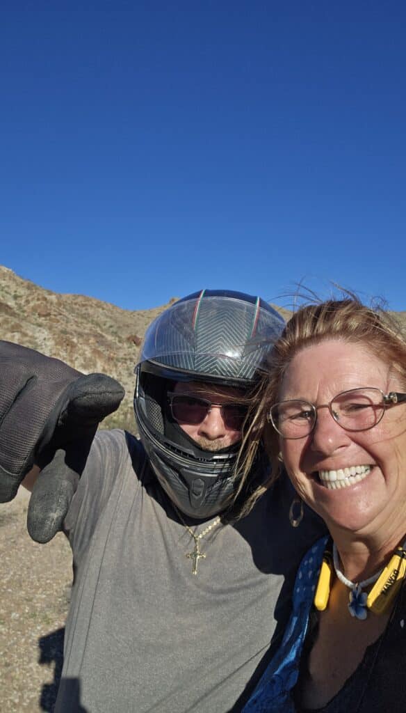 Smiling woman snaps selfie with man in helmet by Nevada’s rocky hills; perfect for Las Vegas ATV tours or Colorado River rides.