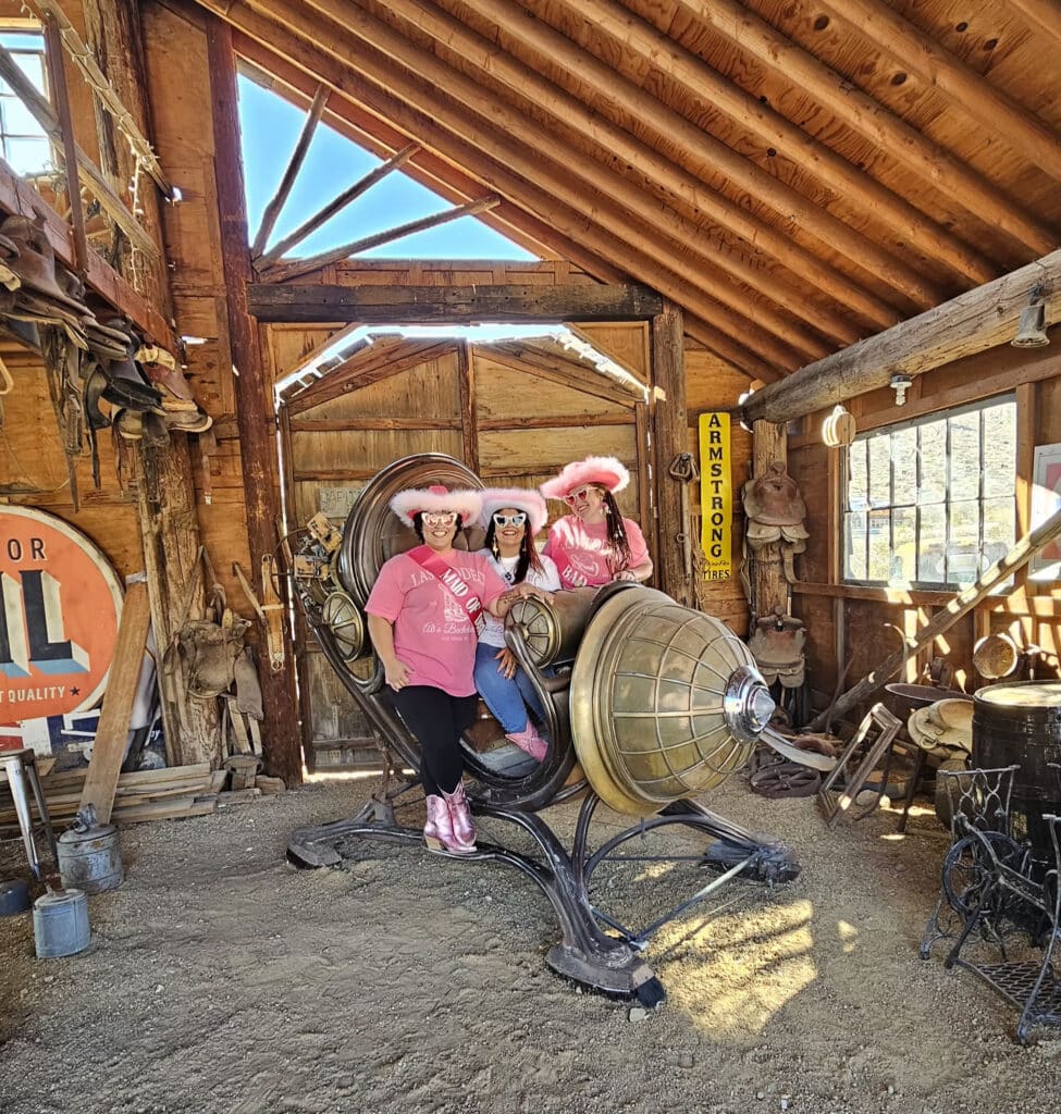 Three women in pink shirts and cowboy hats pose on a steampunk chair in a vintage Nevada barn, perfect for Ghost Town sightseeing.